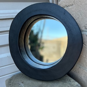 May include: A round black wood framed mirror with a double layered frame. The mirror reflects a blurry image of a building and sky.