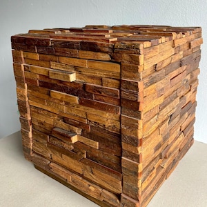 May include: A handcrafted wooden cube with a mosaic-like design. The cube is constructed from various rectangular pieces of wood in shades of brown and tan, creating a textured surface. The cube is a decorative accent piece.