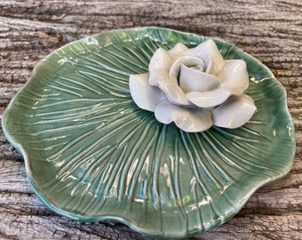 Puede incluir: Un plato de cerámica con forma de hoja de nenúfar verde con una rosa de cerámica blanca en la parte superior.
