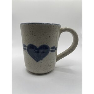 May include: A white ceramic mug with a blue heart design. The mug has a speckled glaze and a handle.
