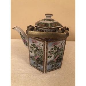 Könnte beinhalten: Eine weiße Porzellan-Teekanne mit sechseckigem Korpus und einem Messinggriff. Die Teekanne ist mit floralen und Vogelmotiven in Rosa-, Grün- und Blautönen verziert. Der Deckel ist ebenfalls mit floralen Motiven verziert.