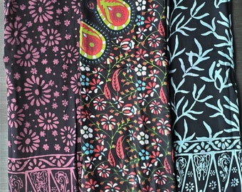 May include: Three black scarves with different patterns. One has pink floral designs, another features paisley and floral motifs with red and green accents, and the third has a light blue leaf pattern. Each scarf has a fringed edge.