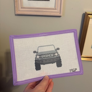 May include: A hand-stitched needlepoint featuring a gray Bronco SUV design. The vehicle is centered on a white canvas, framed by a lavender border. The word "BRONCO" is stitched above the car. The initials "MGP" are in the lower right corner.