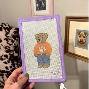 May include: A rectangular needlepoint canvas featuring a brown teddy bear wearing an orange sweater with a white paw print and blue jeans. The canvas is finished with a lavender fabric border and includes the embroidered initials MGP.
