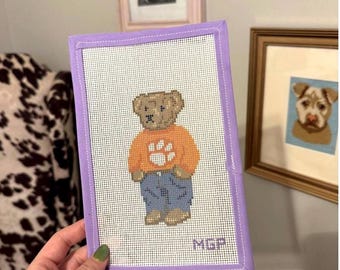 Può includere: Una tela rettangolare per ricamo raffigurante un orsacchiotto marrone che indossa un maglione arancione con un'impronta bianca e jeans blu. La tela è rifinita con un bordo in tessuto color lavanda e include le iniziali ricamate MGP.