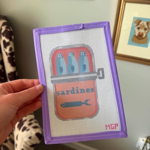 May include: Needlepoint art featuring a can of sardines. The can is salmon-colored with the word "sardines" and a fish illustration. Three blue fish are inside the can. The needlepoint is framed in a lavender border and has the initials "MGP".