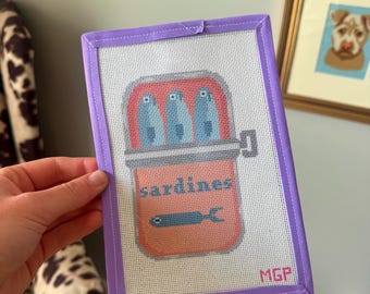 Può includere: Opera d'arte a punto croce raffigurante una scatola di sardine. La scatola è color salmone con la scritta "sardines" e un'illustrazione di pesce. Tre pesci blu sono all'interno della scatola. Il punto croce è incorniciato in un bordo lavanda e ha le iniziali "MGP".