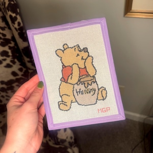 May include: A rectangular needlepoint canvas featuring a yellow bear wearing a red shirt, sitting and holding a tan honey pot labeled Hunny in black script. The canvas is finished with a light purple fabric border and includes the initials MGP.