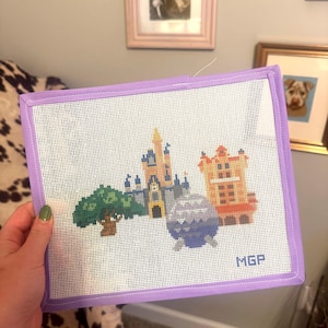 May include: A needlepoint canvas featuring a colorful cityscape design with a castle, tree, and buildings. The canvas is framed in a lavender border and has the initials "MGP" in the corner.