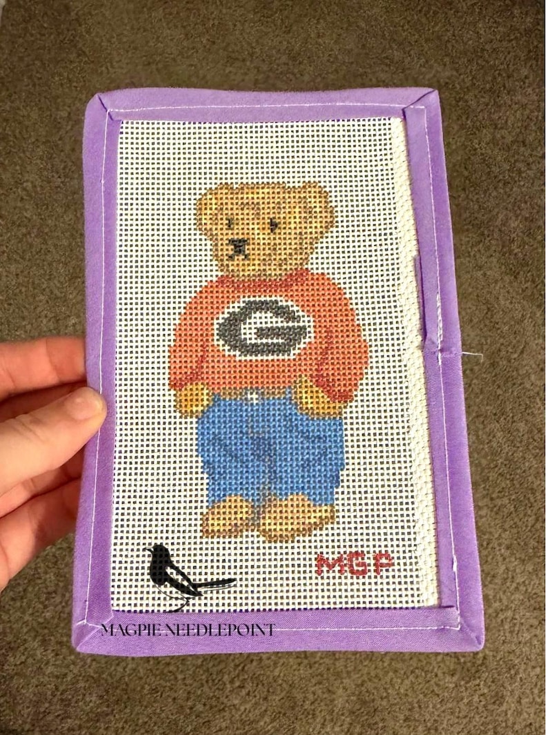 May include: A rectangular needlepoint canvas featuring a tan teddy bear wearing an orange sweater with a black letter G and blue pants. A small black bird is at the bottom left, and the red text MGP is at the bottom right. The canvas has a purple fabric border.