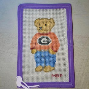 May include: A rectangular needlepoint canvas featuring a tan teddy bear wearing an orange sweater with a black letter G and blue pants. The canvas is bordered by a purple fabric binding and includes the red embroidered initials MGP.