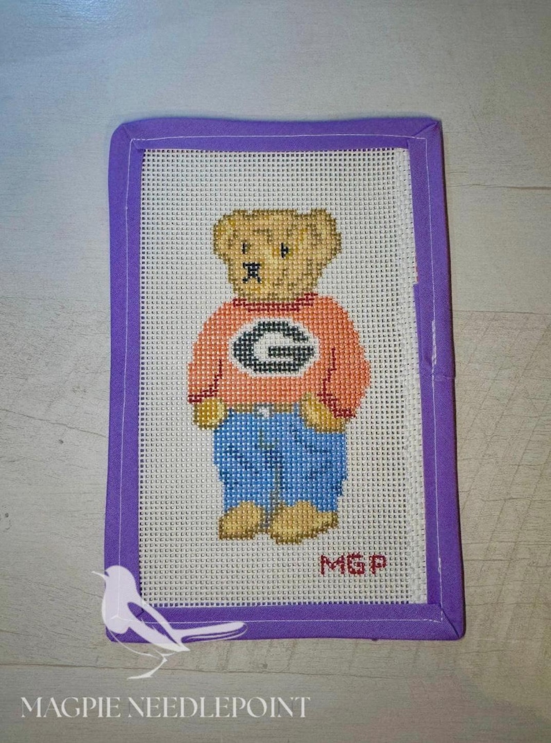 May include: A rectangular needlepoint canvas featuring a tan teddy bear wearing an orange sweater with a black letter G and blue pants. The canvas is bordered by a purple fabric binding and includes the red embroidered initials MGP.