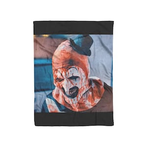 May include: A blanket featuring a close-up of a scary clown with a red and white face, black makeup, and a small black hat. The clown's face is covered in red blood-like paint. The background is a light blue and white.