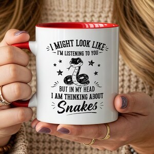May include: A white ceramic mug with a red interior and handle. The front features a black graphic of a snake surrounded by stars and the text: I MIGHT LOOK LIKE I'M LISTENING TO YOU BUT IN MY HEAD I AM THINKING ABOUT Snakes.