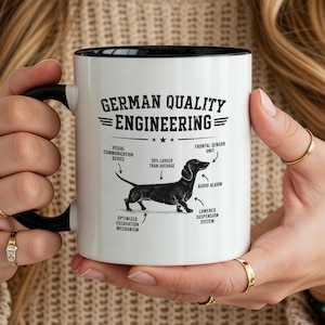 May include: A white ceramic mug with a black handle and interior rim. The mug features a black graphic of a dachshund with text labels reading GERMAN QUALITY ENGINEERING, VISUAL COMMUNICATION DEVICE, 50% LARGER THAN AVERAGE, FRONTAL SENSOR UNIT, AUDIO ALARM, and OPTIMIZED EXCAVATION MECHANISM.