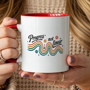 May include: A white ceramic mug with a red interior and handle. The front features the black text 'Progress is not linear' accompanied by colorful, wavy, retro-style lines in orange, yellow, green, and blue, accented with small flower icons.