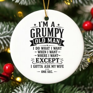 May include: A round white ceramic ornament featuring black text that reads: I'M A GRUMPY OLD MAN I DO WHAT I WANT WHEN I WANT WHERE I WANT EXCEPT I GOTTA ASK MY WIFE ONE SEC. The ornament is suspended by a thin gold string at the top.