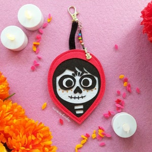 Könnte beinhalten: Ein roter und schwarzer Filz-Schlüsselanhänger mit einem stilisierten Totenkopf-Design und einem kleinen Gitarrenanhänger. Der Schlüsselanhänger ist mit bunten Perlen und einem goldfarbenen Verschluss verziert. Der Hintergrund umfasst orangefarbene Ringelblumen, rote Nelken und kleine Kerzen.