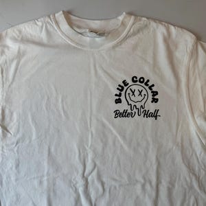 Puede incluir: Una camiseta blanca de manga corta con un gr&aacute;fico negro en el pecho. El dise&ntilde;o incluye una cara sonriente que se derrite con ojos en forma de X, rodeada por el texto "BLUE COLLAR" arriba y el texto en cursiva "Better Half" debajo.