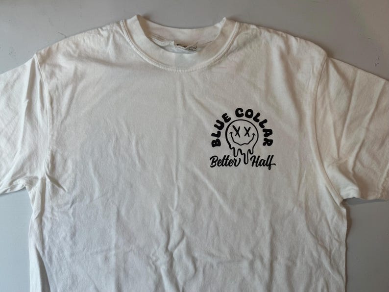 Puede incluir: Una camiseta blanca de manga corta con un gr&aacute;fico negro en el pecho. El dise&ntilde;o incluye una cara sonriente que se derrite con ojos en forma de X, rodeada por el texto "BLUE COLLAR" arriba y el texto en cursiva "Better Half" debajo.