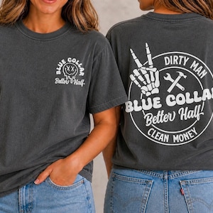 Puede incluir: Una camiseta gris carb&oacute;n con un peque&ntilde;o gr&aacute;fico blanco en el pecho que muestra una cara sonriente y el texto Blue Collar Better Half. La parte posterior presenta un gran gr&aacute;fico circular blanco con una mano esquel&eacute;tica, herramientas y el texto Dirty Man.