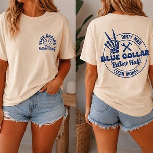 Puede incluir: Una camiseta de manga corta color crema con un peque&ntilde;o gr&aacute;fico azul en el pecho que muestra una cara sonriente derriti&eacute;ndose y el texto Blue Collar Better Half. La parte trasera muestra un gran gr&aacute;fico circular azul con una mano esquel&eacute;tica, herramientas y texto.