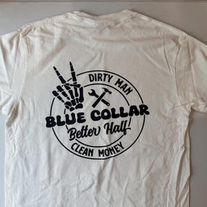 Puede incluir: Una camiseta blanca de manga corta con un gr&aacute;fico circular negro en la parte posterior. El dise&ntilde;o incluye una mano de esqueleto haciendo el signo de la paz, una llave inglesa y un martillo, y el texto: DIRTY MAN, BLUE COLLAR, Better Half!, CLEAN MONEY.