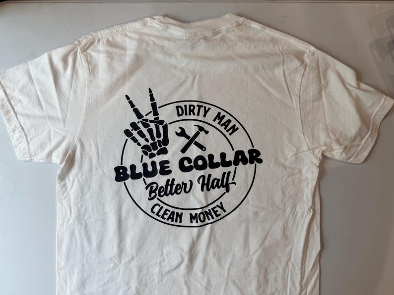 Puede incluir: Una camiseta blanca de manga corta con un gr&aacute;fico circular negro en la parte posterior. El dise&ntilde;o incluye una mano de esqueleto haciendo el signo de la paz, una llave inglesa y un martillo, y el texto: DIRTY MAN, BLUE COLLAR, Better Half!, CLEAN MONEY.