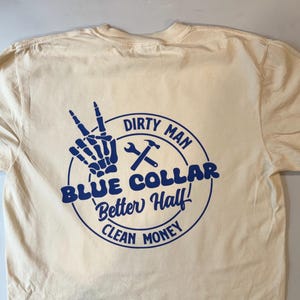Puede incluir: Una camiseta de color crema con un gr&aacute;fico azul en la parte posterior. El dise&ntilde;o incluye una mano de esqueleto haciendo el signo de la paz, una llave inglesa, un martillo y el texto: DIRTY MAN, BLUE COLLAR, Better Half!, CLEAN MONEY, en un c&iacute;rculo.