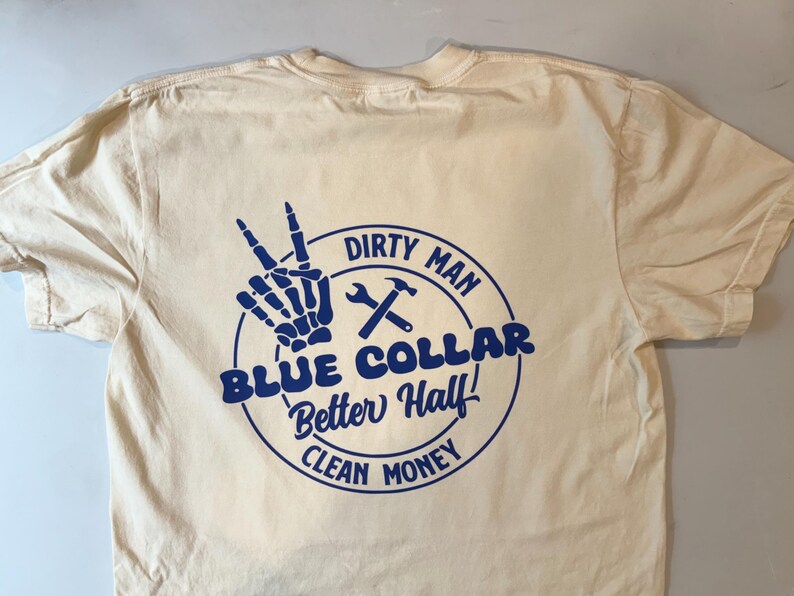Puede incluir: Una camiseta de color crema con un gr&aacute;fico azul en la parte posterior. El dise&ntilde;o incluye una mano de esqueleto haciendo el signo de la paz, una llave inglesa, un martillo y el texto: DIRTY MAN, BLUE COLLAR, Better Half!, CLEAN MONEY, en un c&iacute;rculo.