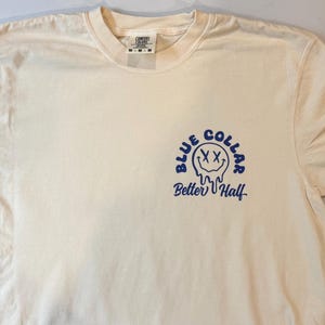Puede incluir: Una camiseta de manga corta de color crema con un gr&aacute;fico azul en el pecho izquierdo. El gr&aacute;fico muestra una cara sonriente que se derrite con ojos en forma de X, rodeada por el texto "BLUE COLLAR" y las palabras "Better Half" debajo.