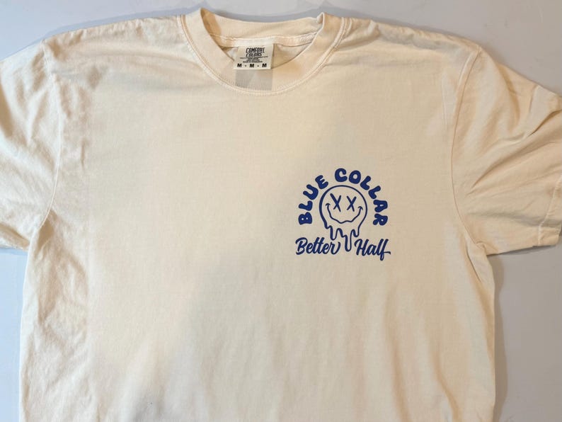 Puede incluir: Una camiseta de manga corta de color crema con un gr&aacute;fico azul en el pecho izquierdo. El gr&aacute;fico muestra una cara sonriente que se derrite con ojos en forma de X, rodeada por el texto "BLUE COLLAR" y las palabras "Better Half" debajo.