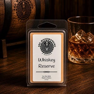 May include: A clear plastic package containing a rectangular soy wax melt labeled "Whiskey Reserve." The label includes the text "ARG Woodworking Designs" and a logo. A glass of whiskey with ice cubes sits to the right. A wooden barrel is in the background.