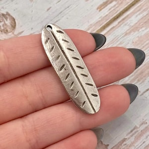 Peut inclure: Un charm en forme de plume en métal argenté, avec une surface texturée et un petit trou en haut. Le motif de la plume présente une veine centrale avec des lignes diagonales de chaque côté. Idéal pour la création de bijoux ou les projets d'artisanat.