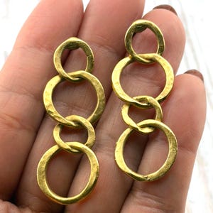 May include: Gold-toned, linked circle earrings. Each earring features three interconnected, hammered-texture rings. The earrings have a warm, metallic finish, and are designed to dangle. The jewelry is shown against a neutral background.