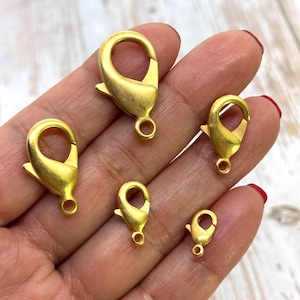 May include: Five gold-toned metal lobster clasps of varying sizes. The clasps are shown on a hand.