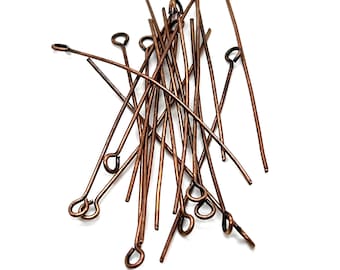 May include: A collection of copper-toned metal eye pins, each featuring a straight wire shaft and a small circular loop at one end. These jewelry findings are arranged in a loose, overlapping pile, showcasing the metallic finish and wire structure.