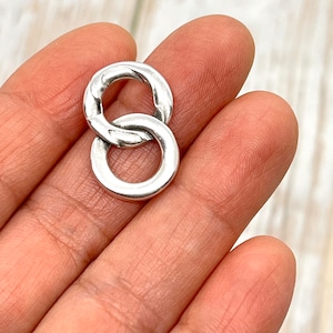 May include: A silver-colored, interlocking double ring charm. The two rings are connected, forming a figure eight shape. The charm has a slightly textured surface and is held in a hand.