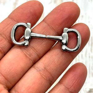 May include: A silver-colored horse bit, a piece of equestrian equipment, is held in a hand. The bit has a central bar connecting two D-shaped rings. The metal has a shiny, reflective surface.