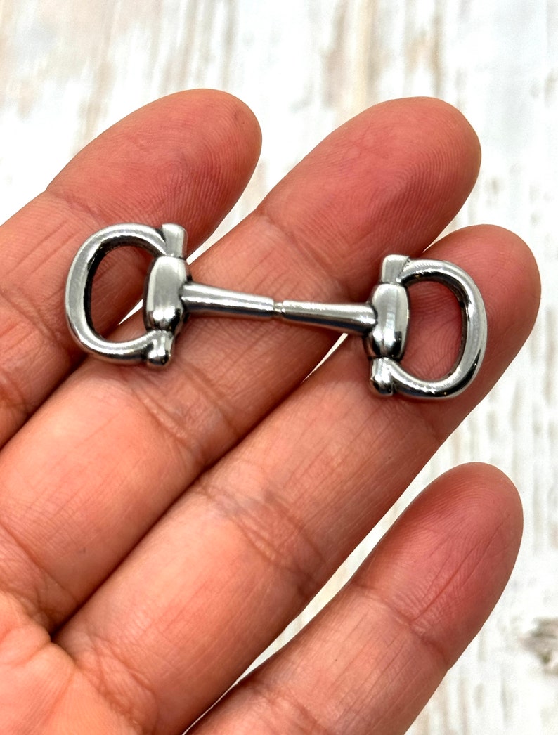 May include: A silver-colored horse bit, a piece of equestrian equipment, is held in a hand. The bit has a central bar connecting two D-shaped rings. The metal has a shiny, reflective surface.
