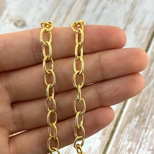 May include: Two strands of gold-colored chain with oval links. The links have a textured, rope-like design. The chains are held against a light-colored wooden surface.