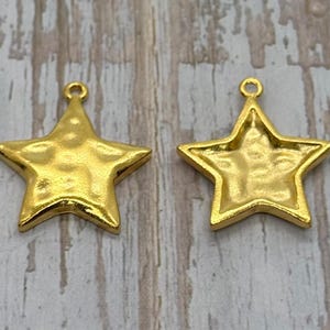 May include: Two gold-colored star-shaped charms. One charm is solid, while the other has a hollowed-out back. Both have a textured surface and a small loop at the top for hanging. The charms are on a light wood-grain background.