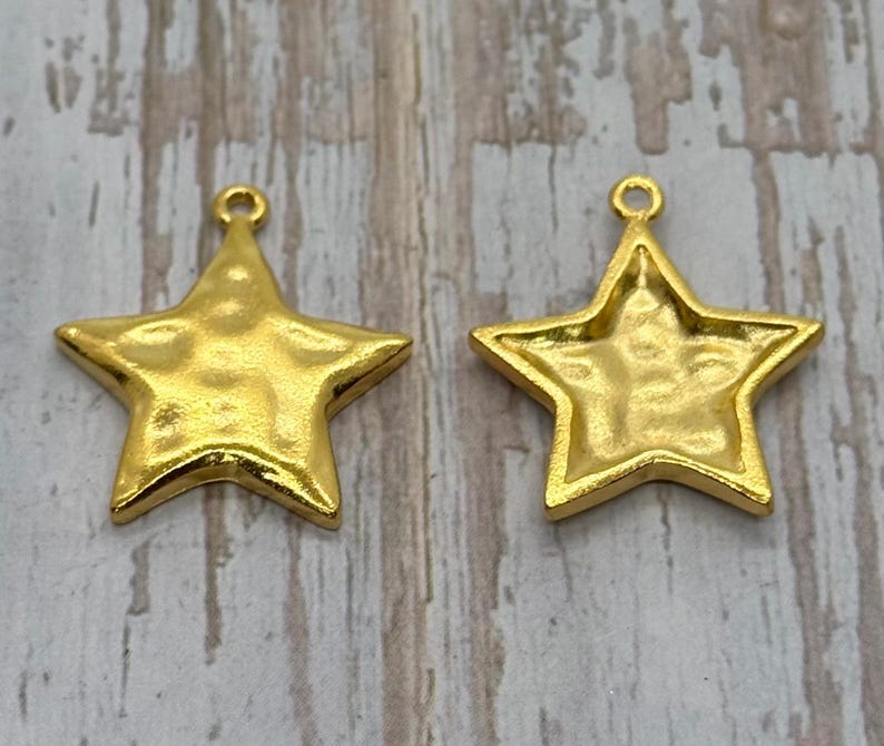 May include: Two gold-colored star-shaped charms. One charm is solid, while the other has a hollowed-out back. Both have a textured surface and a small loop at the top for hanging. The charms are on a light wood-grain background.