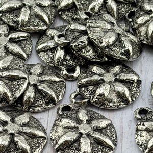 Op de afbeelding: Een verzameling zilverkleurige metalen knopen. Elke knoop heeft een bloemenpatroon met gedetailleerde texturen en een lus om aan te naaien. De knopen zijn rond en lijken van vintage of antiek materiaal te zijn.