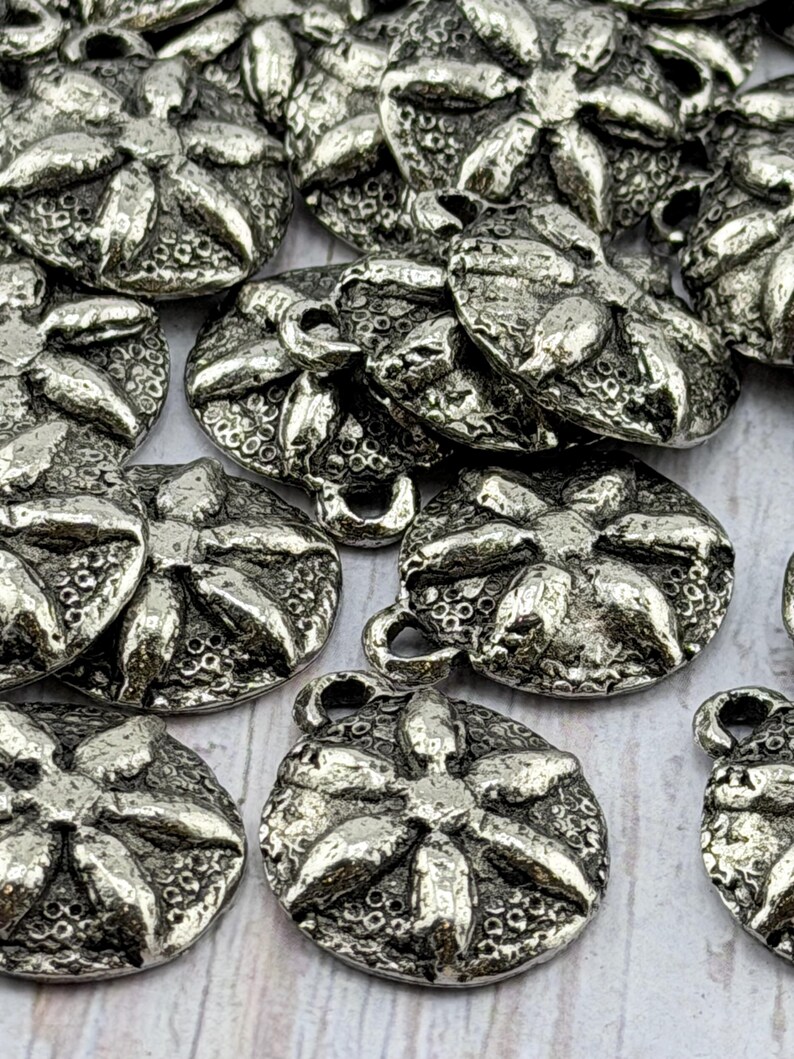 Op de afbeelding: Een verzameling zilverkleurige metalen knopen. Elke knoop heeft een bloemenpatroon met gedetailleerde texturen en een lus om aan te naaien. De knopen zijn rond en lijken van vintage of antiek materiaal te zijn.