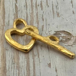 May include: A gold-colored metal charm in the shape of a heart with a key. The heart-shaped charm has a textured surface and a loop at the top. The key is attached to the heart and has a similar textured finish.