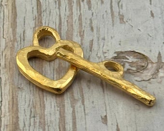 May include: A gold-colored metal charm in the shape of a heart with a key. The heart-shaped charm has a textured surface and a loop at the top. The key is attached to the heart and has a similar textured finish.