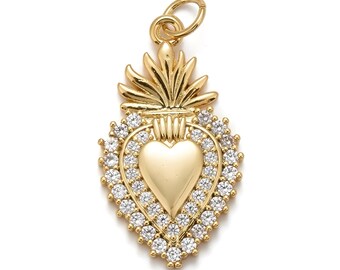 May include: Gold-colored pendant with a heart-shaped center, surrounded by a halo of small, clear gemstones. The pendant has a decorative top with a loop for a chain. The design is symmetrical and detailed.