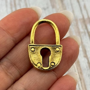 May include: A small, gold-colored padlock with a rounded top and a keyhole in the center. The padlock has a vintage or antique appearance, with a slightly worn texture. The metal has a warm, brassy tone.