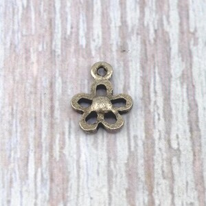 May include: A small, antique bronze-colored flower charm with five petals and a loop at the top for stringing. The charm has a slightly textured surface and is designed for jewelry making or crafting.